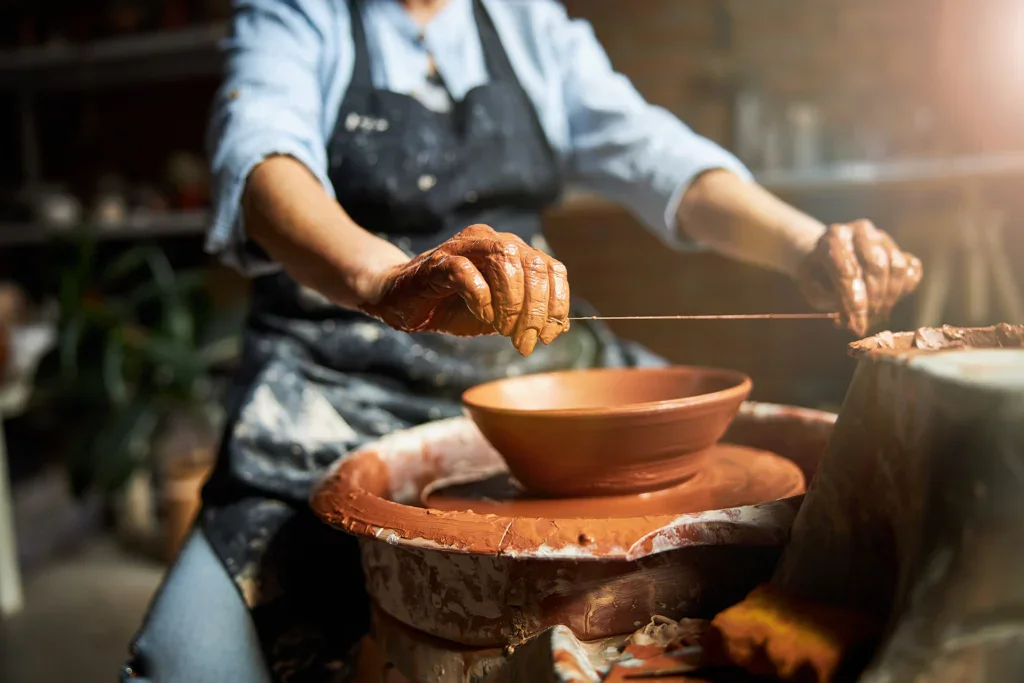 Pottenbakken benodigdheden: Een vrouw in haar keramiek atelier aan de draaischijf terwijl ze met een draad een gedraaide kom van de draaischijf afsnijdt