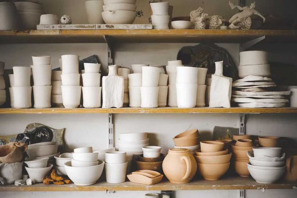 keramiek vragen: verschillende legplanken in een klei atelier gevuld met stapels biscuit gebakken kommen, bekers, theepotten en flessen.