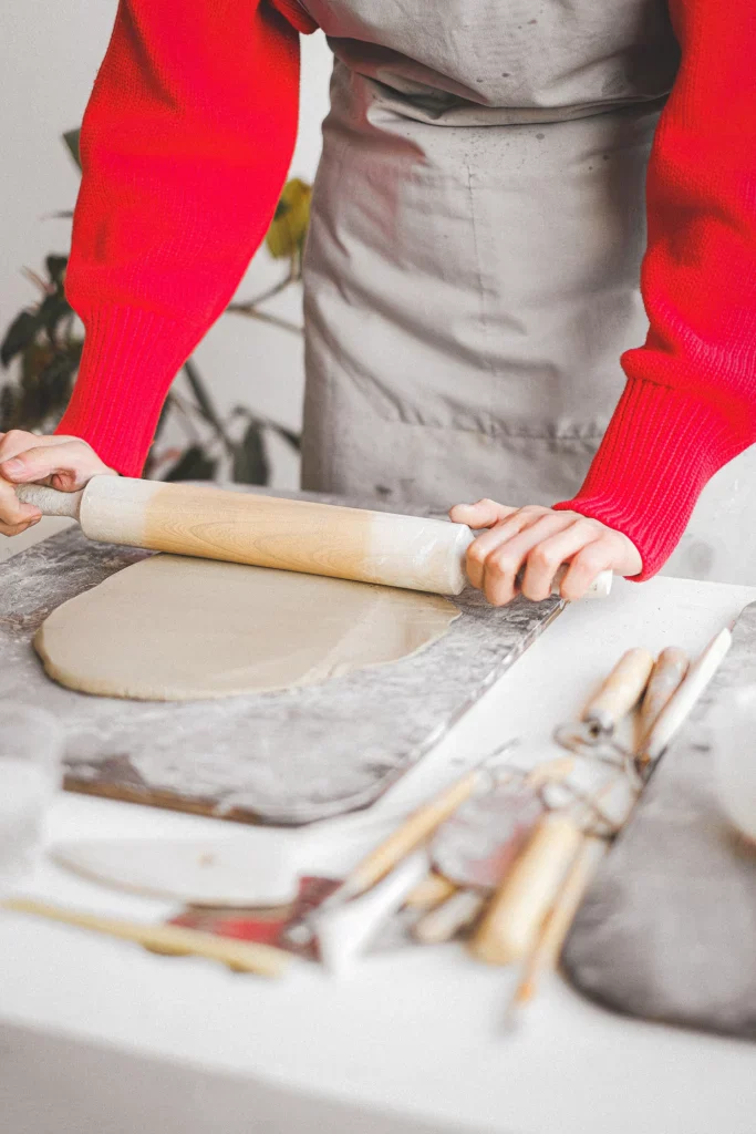 Iemand rolt klei uit tot kleirollen op een kleiplaat met op tafel verschillende instrumenten om klei te bewerken
